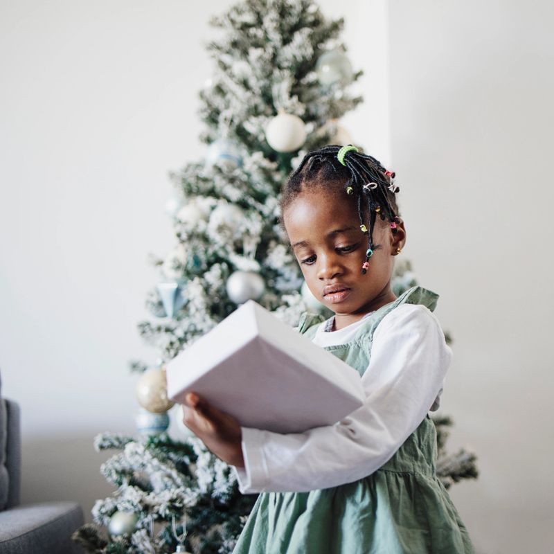 Little African American girl in dress standing in front of the Christmas tree and holding wrapped gift in hands