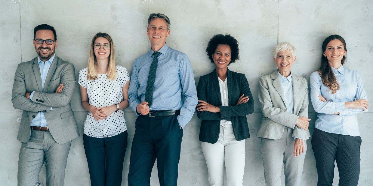 Midlife women and men in workplace attire pose before a grey stone wall and smile