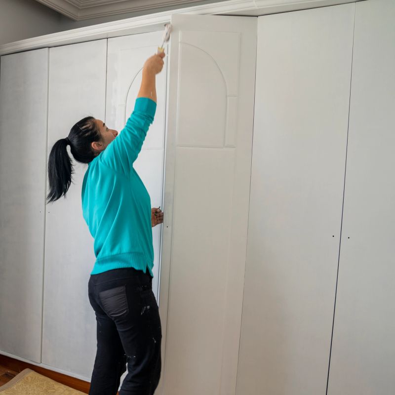 Photo of mature adult woman painting wardrobe at home. She is wearing an orange sweater and a bandana. The paint is white. Shot indoor  with a full frame mirrorless camera.