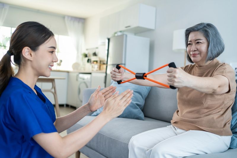 Asian Disabled old woman doing physiotherapist with support from nurse. Senior elder handicapped female sit on sofa in living room use resistance stretch band exercise for patient in home nursing care