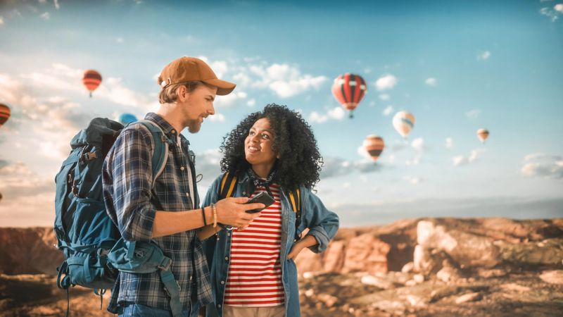 Young Diverse Couple of Hikers are Checking Tourist Information and Directions on Their Smartphone on Top of Rocky Canyon Valley. Backpackers on Adventure. Hot Air Balloon Festival in National Park.
