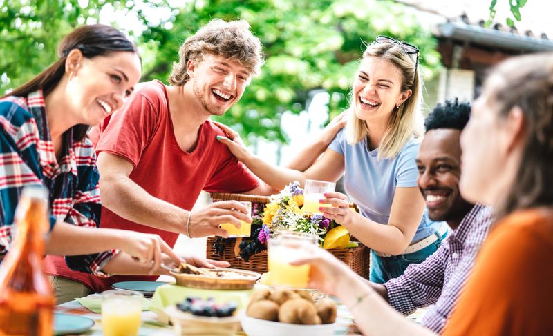 Happy young men and women toasting healthy orange fruit juice at farm house picnic - Life style concept with alternative friends having fun together on afternoon relax time - Bright vivid filter