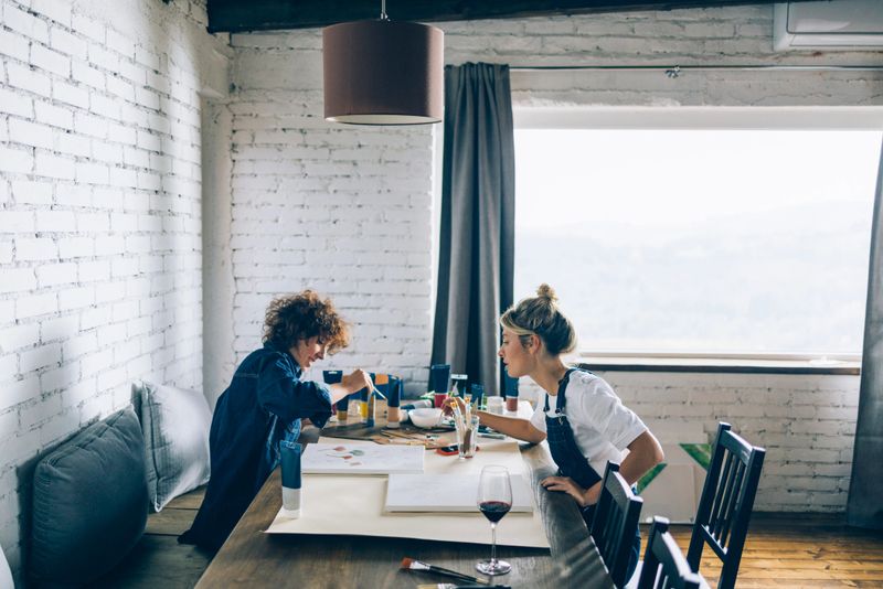 Two smiling friends sitting at table and painting with watercolors
