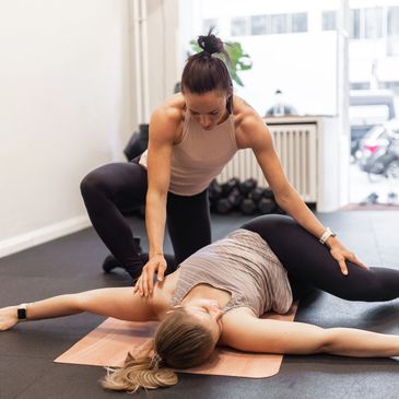 A trainer assists a woman stretching on a mat in a gym.