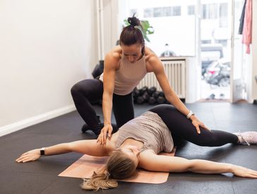 A trainer assists a woman stretching on a mat in a gym.