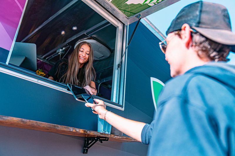 Young Female Business Owner Standing Giving Credit Card Reader to a Customer to Use a Smartphone to Pay for Goods Using NFC Cashless Technology at a Food Truck Concession Stand