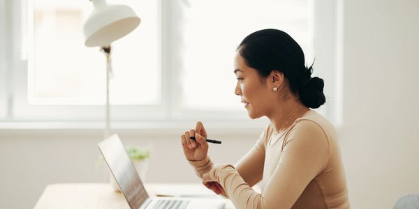 Woman working on laptop in a bright room, holding a pen.