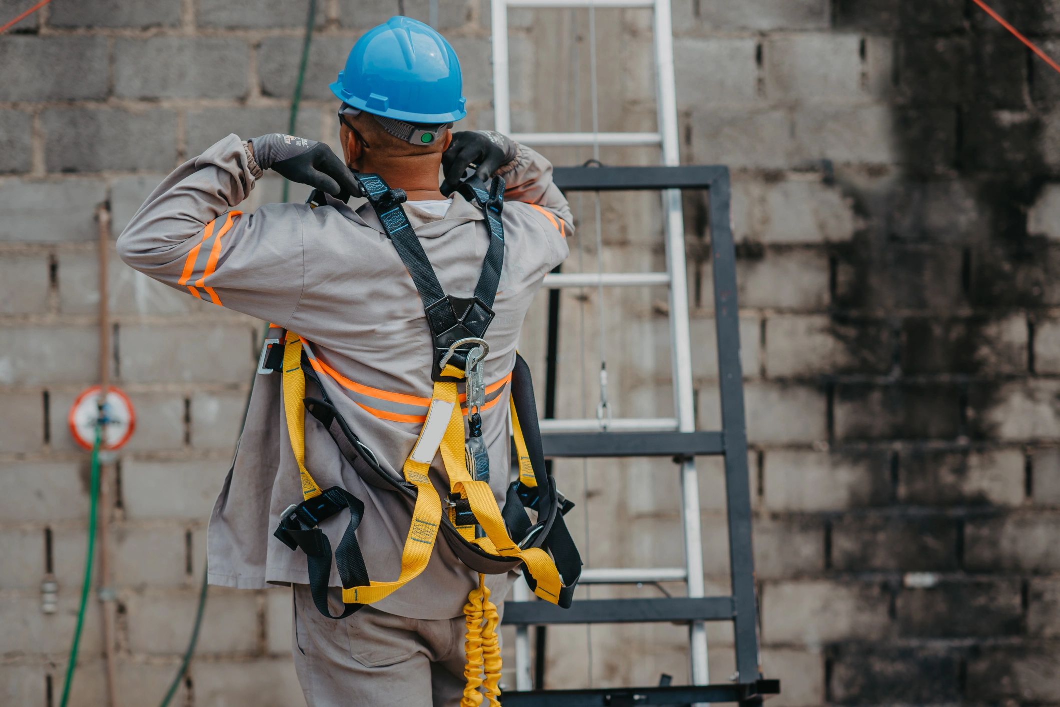 Construction worker in a hard hat putting on a full-body fall protection harness next to a ladder