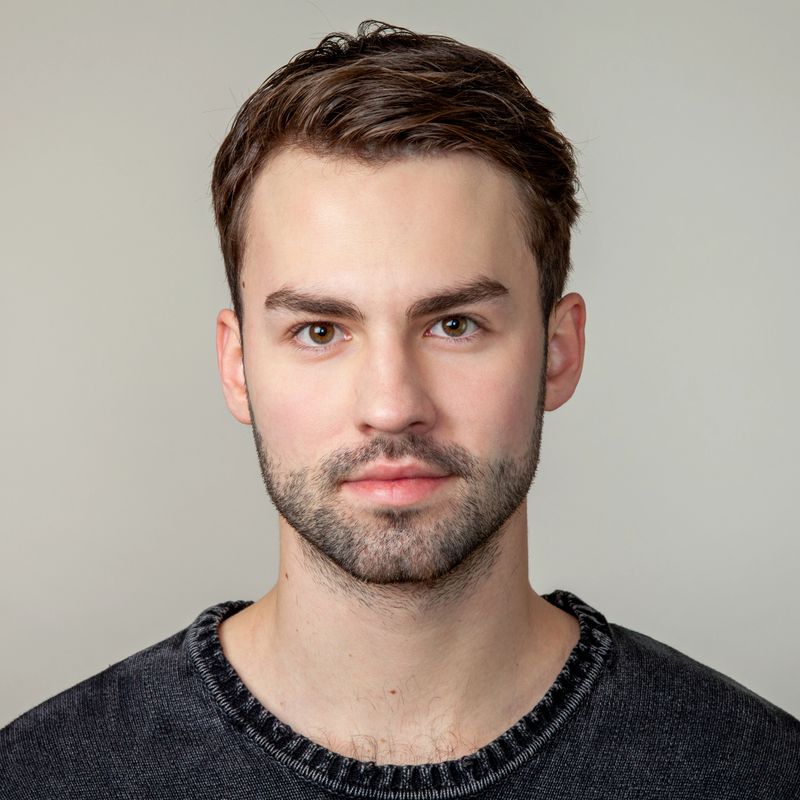 Close up studio portrait of attractive 20 year old bearded man with brown hair in black sweater on gray background