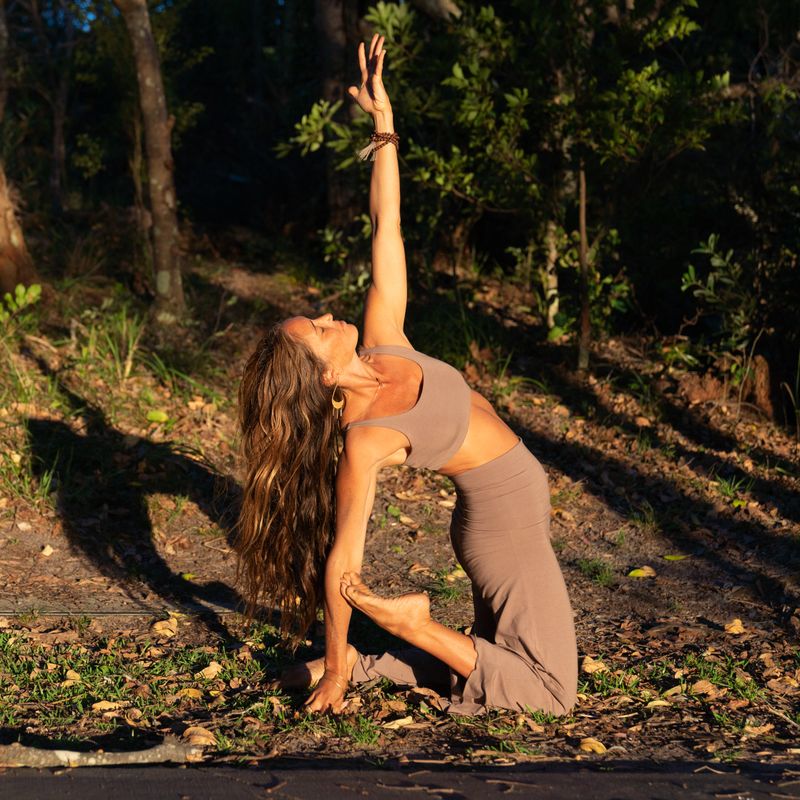 Full length shot of a woman in nature doing a back band Yoga pose in the golden afternoon sun, she is kneeling with one arm up and one down, she is looking up and her hair is flowing,  she is wearing a tan top and tan flare pants, surrounded by trees in nature