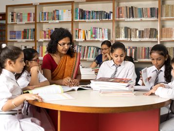 Teacher guiding students in a library around a table with books.