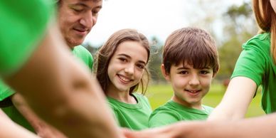 Smiling group in green shirts stacking hands in teamwork gesture outdoors.