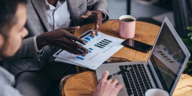 Two men discussing business charts and working on a laptop at a wooden table.