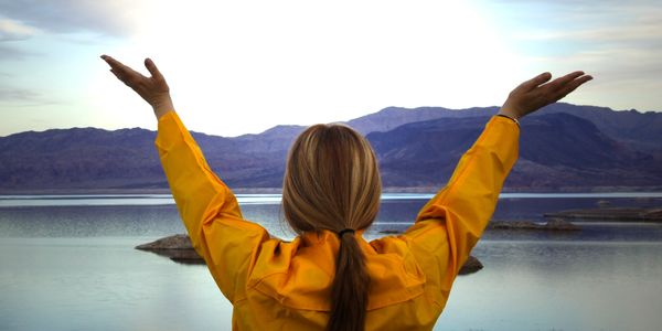 Person in yellow jacket raising arms by a serene lake with mountains in the background.