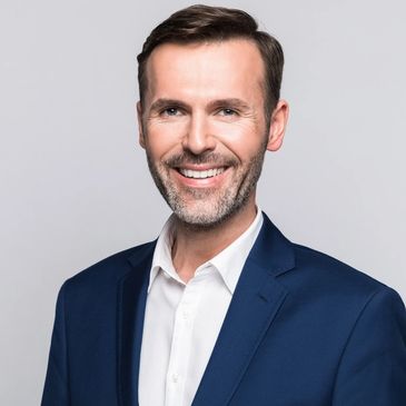 Smiling man in a navy suit and white shirt against a gray background.