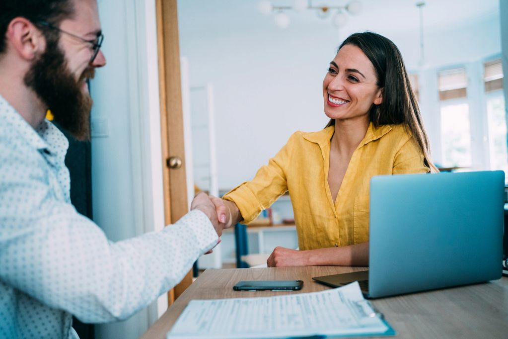 Two people shaking hands across a desk with a laptop and documents.