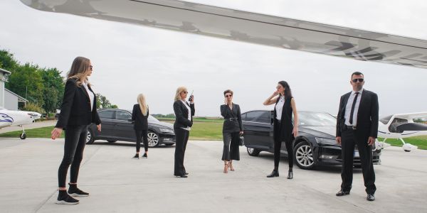 A group of six people in business attire near an airplane and cars.