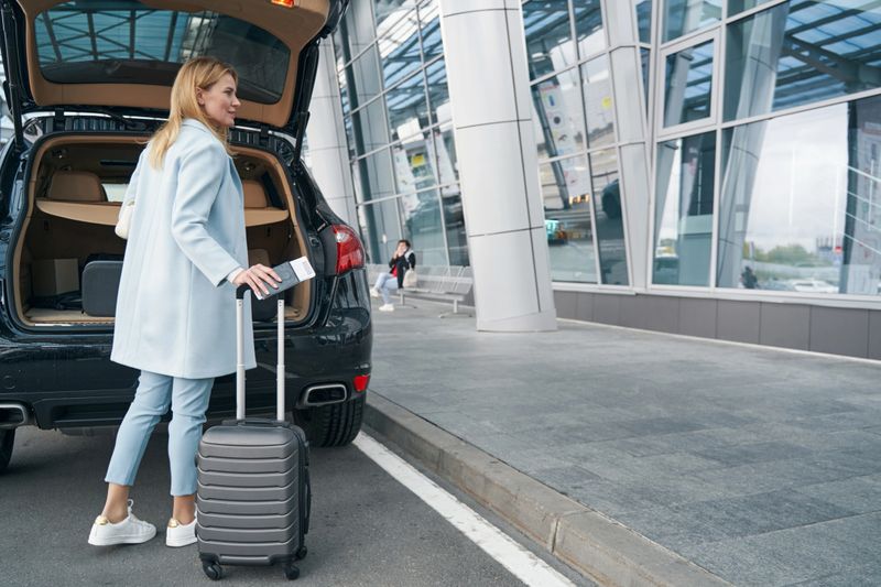 Back view of woman with boarding ticket and trolley suitcase standing at rear of vehicle