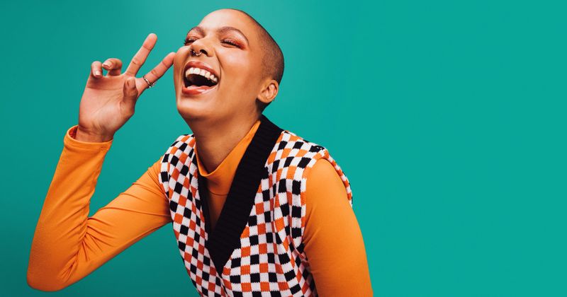 Carefree young woman showing the peace sign in a studio. Happy young woman laughing with her eyes closed while standing against a turquoise background. Woman with septum ring feeling cheerful.
