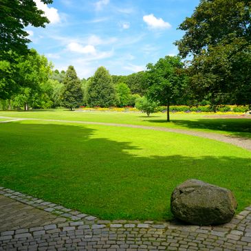 A sunny park with green grass, trees, and a stone on a paved path.
