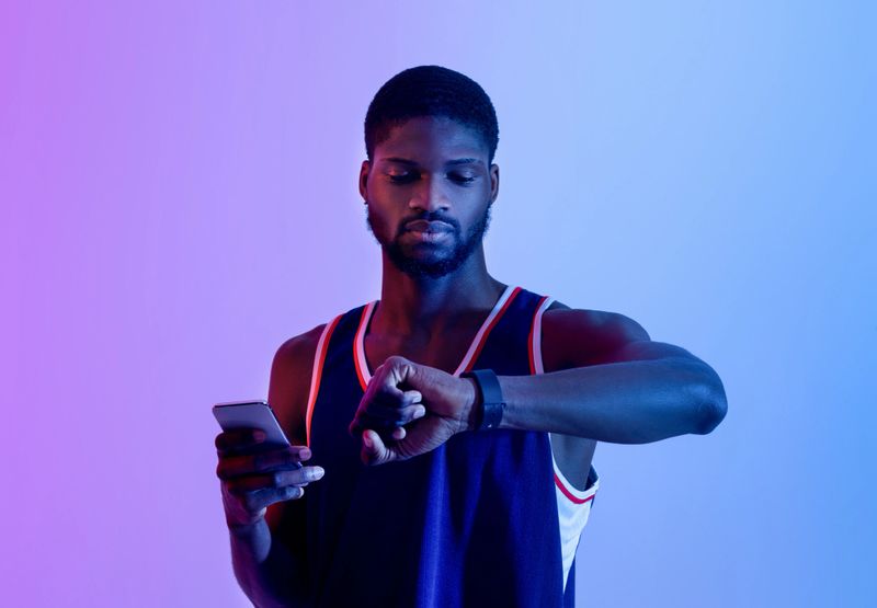 Athletic black man synchronizing his smartwatch with cellphone before training in neon light. Fit young guy checking fitness tracker, using mobile device. Modern technologies and active lifestyle