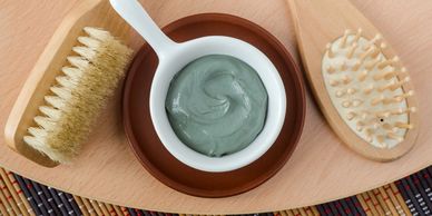 Wooden brushes and a white bowl with green clay mask on a wooden board.