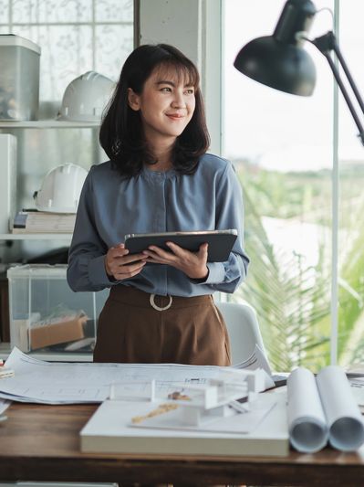 A woman holding a tablet, standing by a desk with architectural plans and helmets.