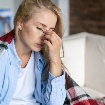ME sufferer sits on sofa with blanket around her shoulders, eyes closed, hand on face