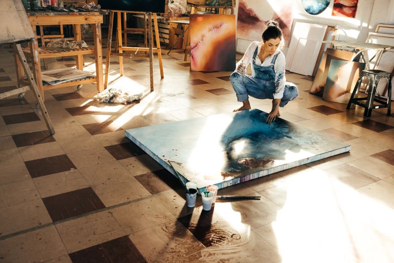 Female painter making a blue painting on a canvas. Creative young woman touching her painting while squatting on the floor in her art studio. Artistic young woman working on a new project.