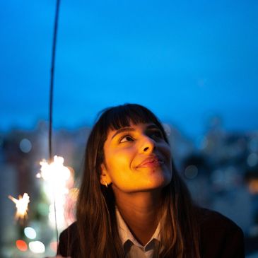 A young woman smiles softly under sparklers at dusk.