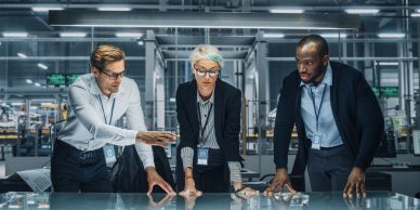 Three professionals collaborating over a transparent digital table in a modern industrial setting.