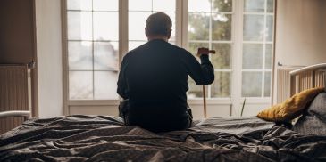 Elderly man sitting on a bed holding a cane, facing a window.