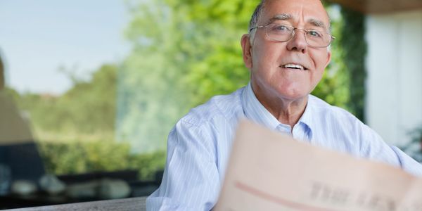 Smiling elderly man reading a newspaper outdoors on a sunny day.