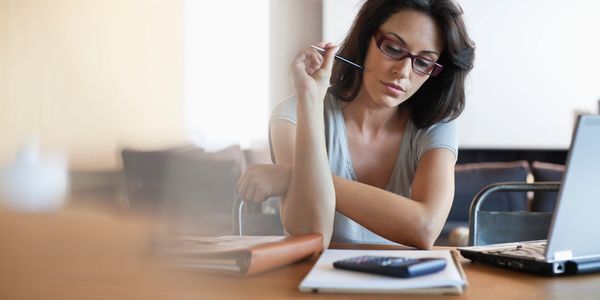 Woman with glasses concentrating on paperwork at a desk with a laptop and calculator.
