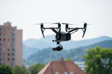 A black drone hovering outdoors with mountains and buildings in the background.