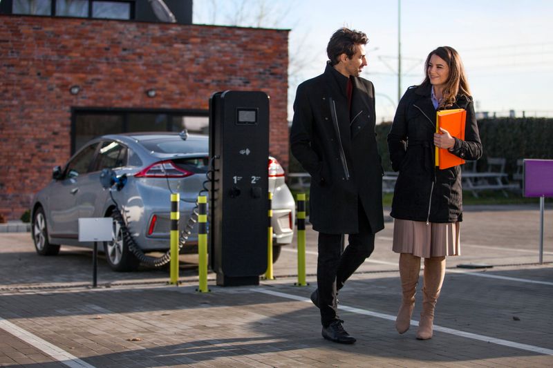 Photo of a young couple or office workers on a parking lot, charging their electric car, she is holding a file folder