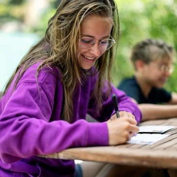 Smiling girl writing outdoors at a wooden table.