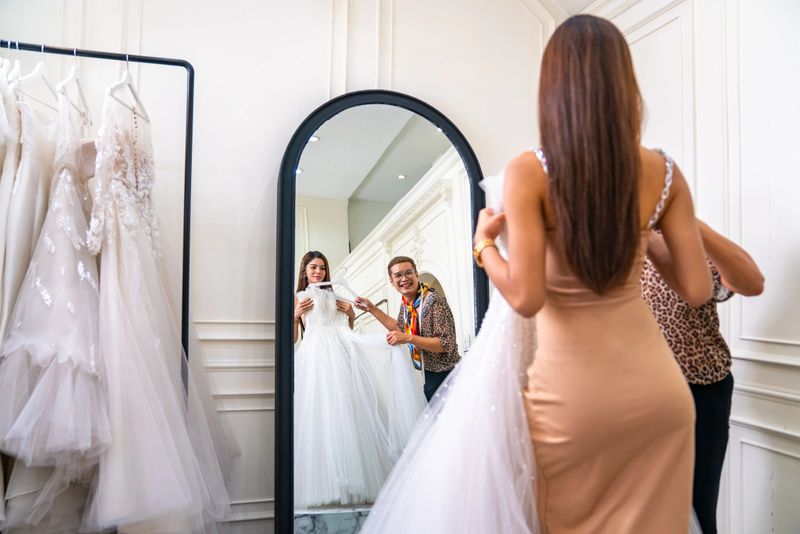 Asian LGBTQ guy bridal shop owner helping female bride customer choosing and trying on wedding gown at wedding studio. Small business entrepreneur wedding planner and marriage ceremony concept