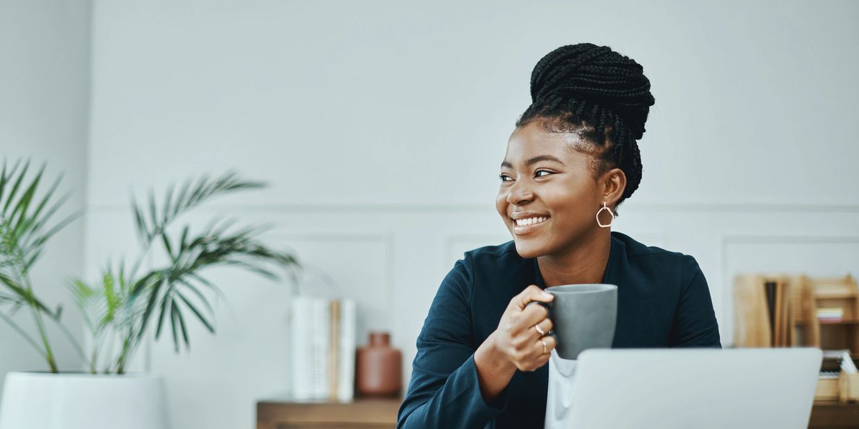 Smiling woman with braided hair holding a mug while sitting at a desk.