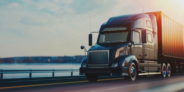 A black semi-truck driving on a highway near a body of water at sunset.