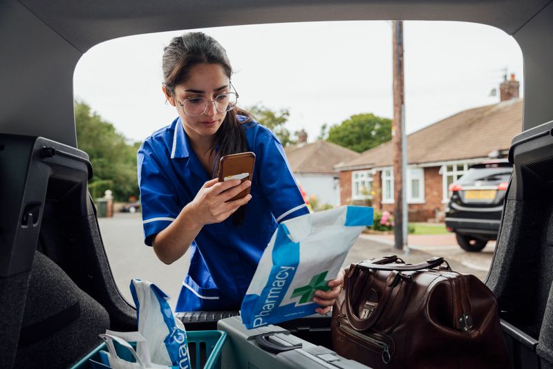 Nurse arriving to a patients home in the North East of England getting her bags and prescriptions needed from the boot of her car, dressed in her uniform. She is taking a photo on her mobile phone of a prescription bag.