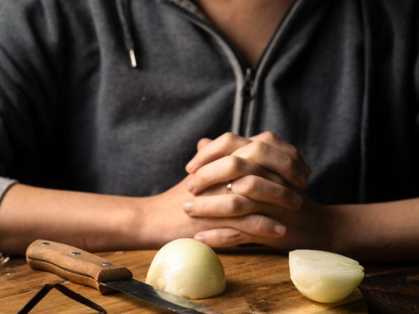 Person sitting with folded hands near a halved onion and knife on a wooden board.