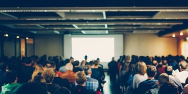 Image of large conference room full of people with a presenter in front of a screen  