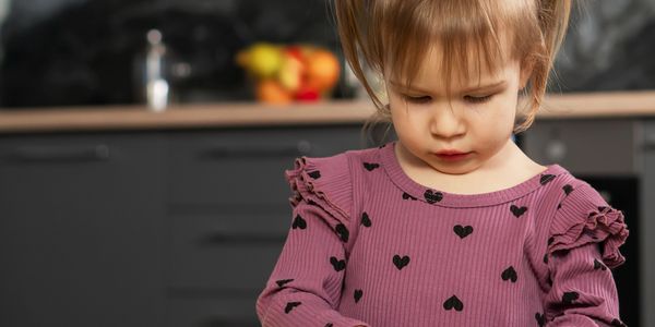 A young girl in a heart-patterned dress looks down thoughtfully in a kitchen.