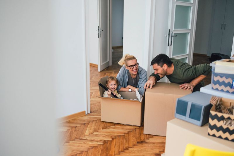 Family of three is moving in or out of a room in an apartment or a house. Mother and father are playing with the child on the floor of the room. The child is sitting in a box and holding what seems to be a vacuum cleaner stick. They are all smiling and the father is leaning towards the adorably cute kid.