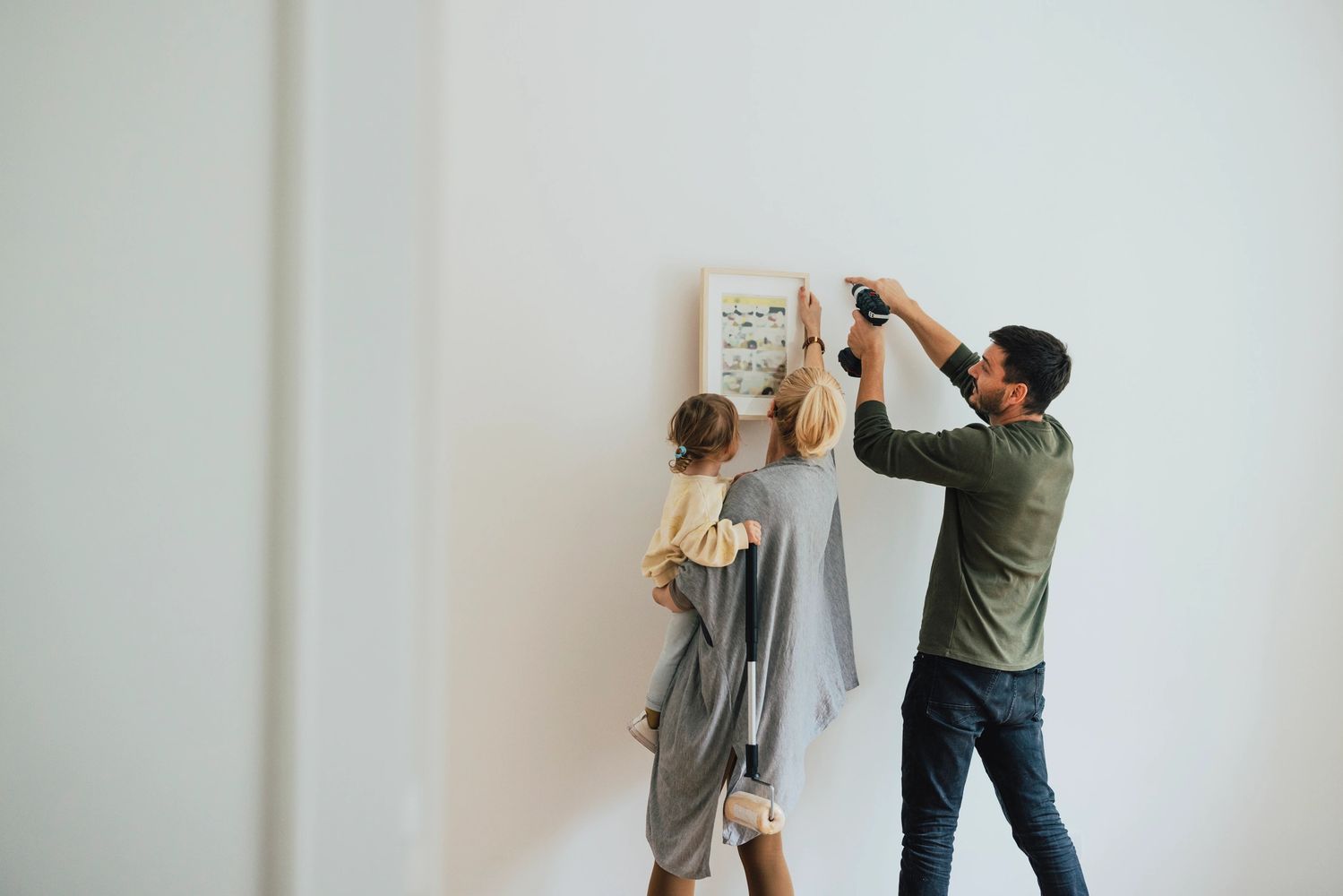 A couple hangs a picture frame on a white wall while holding their child.