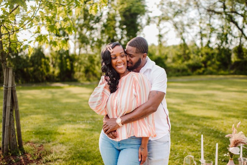 A black couple sits at a private romantic picnic enjoying each others company after a surprise proposal in the park. Happy and elated smiles and interaction relaxing on pillows and a tent.