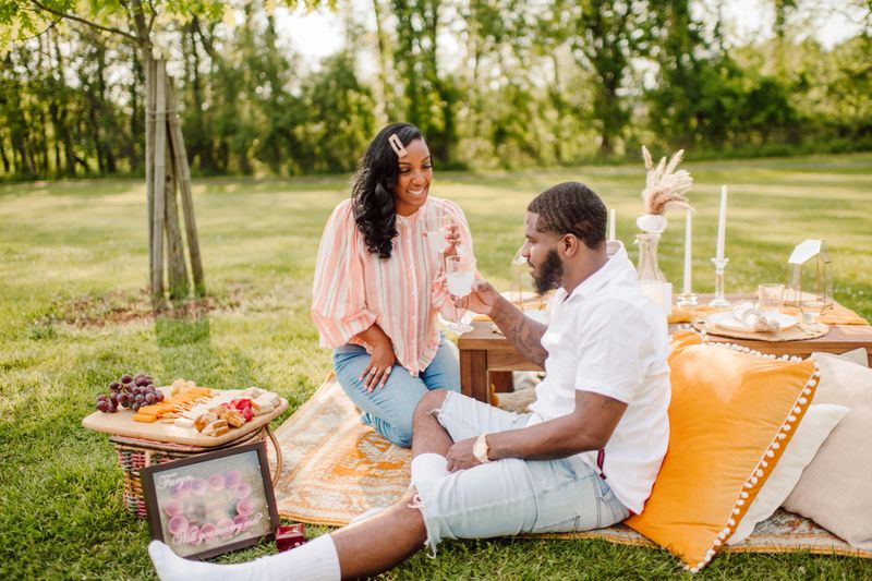 A black couple sits at a private romantic picnic enjoying each others company after a surprise proposal in the park. Happy and elated smiles and interaction relaxing on pillows and a tent.