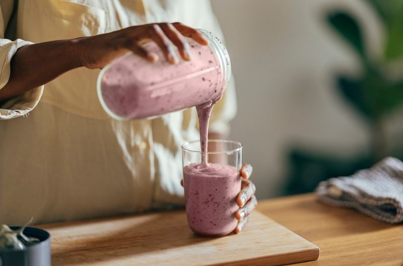 Close-up of hands of an Afro-American woman wearing beige shirt and pouring smoothie into a glass on a kitchen table. There is a part of a blender, a cutting board and a cloth on the desk in front of her. In the corner of a room sits a big plant.