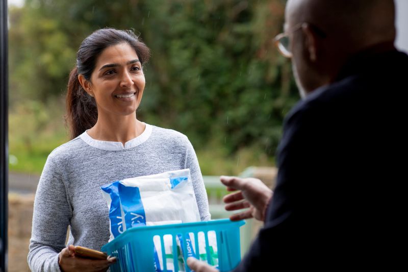 A community care volunteer delivering prescription medicine to a patient at their house. She is standing outside the door and passing it to the patient who is standing inside the house.